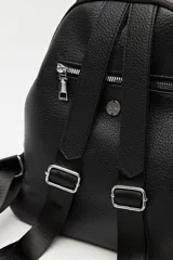 Mochila de símil cuero texturizado color beige, con asa superior, tiras regulables para la espalda y cierre principal metálico. Presenta un detalle trenzado vertical en el frente y un accesorio con flecos y cuenta negra en el lateral.