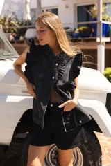 Chaleco de denim lavado negro, sin mangas, con volados estructurados en el cuello y a lo largo del frente. Presenta cierre frontal con botones metálicos plateados y bolsillos laterales.