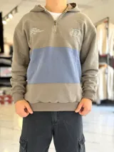 Hoodie de diseño en bloques de color, combinando tono gris y una franja central en celeste. Cuenta con capucha, cuello con cierre corto y bolsillos laterales. Presenta detalles de texto y logo bordados en el pecho.