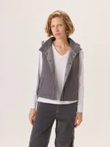 Chaleco gris con cuello alto y capucha, cierre frontal con botones a presión. La parte delantera es de lana gris lisa y la parte interior del cuello y el forro son de tela con patrón de espiga en blanco y negro.