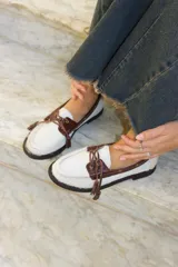 Mocasines de estilo náutico confeccionados en cuero, combinando paneles de cuero con textura de pelo en color blanco y detalles en cuero con textura croco en color marrón. Presentan cordones decorativos con borlas en el empeine y suela plana de goma.