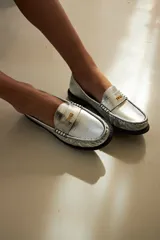Mocasín de cuero con textura efecto cocodrilo en color plateado, con herraje metálico dorado con logo VDM y suela plana oscura.
