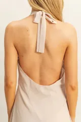 Vestido largo de tela fluida en color beige, con escote halter y detalle de flor aplicada en la cintura. Presenta un diseño asimétrico con caída suave y elegante.