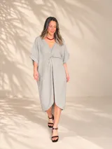 Vestido midi de lino color beige, con diseño oversize, escote en V y detalle de nudo drapeado en el frente. Posee mangas cortas amplias con dobladillo y corte asimétrico, siendo más largo en la parte trasera.