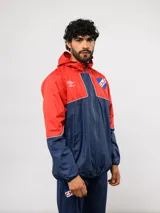 Campera de lluvia Umbro del Club Nacional de Football, con diseño de bloques de color en rojo y azul marino. Presenta capucha, cierre frontal, puños elásticos y detalles de piping blanco. Incluye logo de Umbro y escudo del club en el pecho.