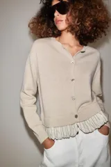 Chaqueta de punto color beige con cuello redondo, manga larga y cierre frontal con botones. Presenta un diseño combinado con volados de tela en el bajo.