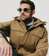 Campera utilitaria de poliéster color tostado, con capucha ajustable, cuello alto, cierre frontal con cremallera y solapa con botones a presión, y bolsillos frontales grandes con solapa.