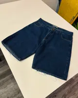 Short de jean de mezclilla azul oscuro, corte ancho y largo (tipo bermuda), con bordes deshilachados y dobladillo sin rematar.