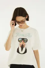 Remera blanca de algodón con cuello redondo y mangas cortas, de corte holgado. En el frente presenta el texto "Farm Rio" en tono celeste y un estampado gráfico de un tucán con anteojos de sol que reflejan un paisaje playero.