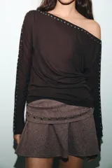 Camiseta de cuello barco y manga larga tipo murciélago, confeccionada en tejido de punto gris melange. Presenta detalle de tachas metálicas a lo largo de los hombros y las mangas.