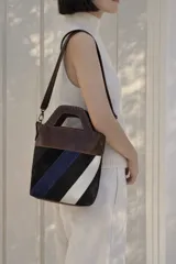 Bolso tipo matera confeccionado en cuero vacuno, con diseño estructurado y manija superior integrada. Presenta un cuerpo con franjas diagonales en gamuza de colores contrastantes (amarillo, verde y azul) sobre una base de cuero marrón, con detalles de costuras artesanales en los bordes.