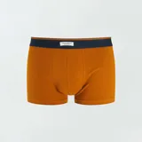 Pack de tres boxers de algodón elástico, uno liso color ocre, uno a rayas horizontales azul y ocre, y uno azul con estampado de hojas color ocre.