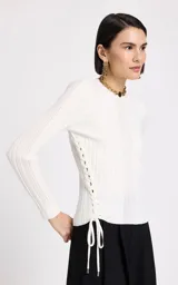 Sweater de punto fino color blanco, con cuello redondo y mangas largas. Presenta una textura acanalada con diseño de puntadas variadas y un detalle lateral de cordones trenzados con cierre de lazo y punteras metálicas.