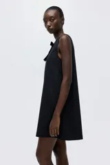 Vestido negro corto sin mangas, de corte evasé, con escote bote y moño decorativo de terciopelo negro en el frente.