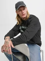 Buzo de manga larga y cuello redondo, color negro, con fit holgado. Presenta un estampado frontal en tonos verde menta y blanco con texto que incluye "Originals Studio", "Daily Basics", "International", "connecting CREATIVITY ACROSS DISTANCES" y "est. 90".