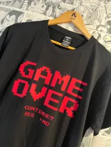 Remera negra de algodón con estampado frontal en rojo que simula tipografía de videojuegos retro, mostrando el texto "GAME OVER" en grande, y debajo "CONTINUE? YES NO".