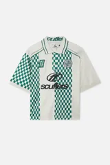 Polo de manga corta color blanco crudo con cuello y ribetes verdes. Presenta un diseño inspirado en camisetas de fútbol con bloques de cuadros verdes y blancos en las mangas y laterales, y una franja central blanca con estampado de logo en negro.