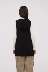 Chaleco largo sin mangas de tejido de punto color negro, con capucha y lazo ajustable en la cintura. Presenta bolsillos frontales y diseño abierto.