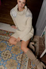 Buzo tipo polera de tejido de punto grueso en color beige, con cuello alto y diseño holgado. Presenta detalles bordados en canutillos plateados con forma de flor en ambos hombros.
