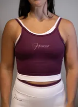 Top deportivo ajustado color morado oscuro con doble capa de tela y tirantes finos blancos. Presenta un logo manuscrito en tono rosado claro en el pecho.