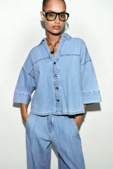 Camisa de denim en tono azul claro, con cuello solapa, escote en pico y cierre frontal con botones. Presenta mangas anchas por debajo del codo y un diseño de bajo asimétrico.