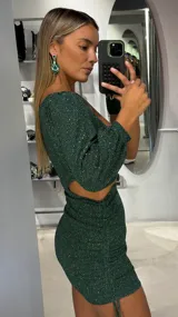 Vestido corto de lurex color verde esmeralda, con diseño de un solo hombro, manga larga abullonada y abertura lateral en la cintura. Presenta un ajuste al cuerpo con detalle fruncido en la falda.