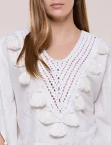 Blusa blanca de silueta holgada con sutil trama en hilo metalizado lurex, escote en V con pechera tejida en crochet y apliques de borlas en el contorno del cuello y las mangas.