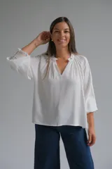 Blusa blanca de corte holgado con cuello redondo fruncido y abertura frontal en V. Presenta mangas tres cuartos con puños elásticos y detalle de tejido texturizado en los mismos.