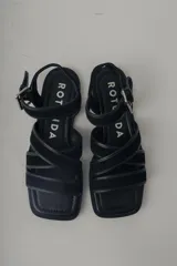 Sandalia plana de cuero negro, con diseño minimalista de tiras finas cruzadas sobre el empeine y una tira ajustable con hebilla metálica en el tobillo.