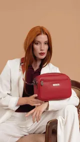 Bandolera de diseño rectangular con textura granulada, color rojo. Cuenta con tres compartimentos principales con cierre, un bolsillo frontal con cierre y placa metálica decorativa con logo. Incluye correa ajustable y extraíble.