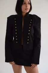 Chaqueta de estilo militar en color negro, con cuello mao y cierre frontal con ganchos decorativos y botones dorados. Presenta hombreras estructuradas y diseño entallado.
