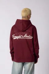 Buzo canguro color bordó con capucha y cordones ajustables. Presenta un bolsillo frontal tipo canguro y un pequeño parche rectangular con diseño gráfico en la manga derecha.