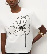 Remera de corte box, levemente amplia, confeccionada en algodón. Presenta cuello redondo, mangas cortas y un diseño de flor bordada en hilo negro en la parte delantera.