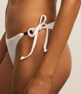 Conjunto de bikini blanco de microfibra acanalada. El corpiño es de estilo triangular con tirantes finos ajustables. La bombacha es de corte clásico con lazos laterales ajustables y detalle de adornos metálicos, diseñada con efecto levanta glúteos.