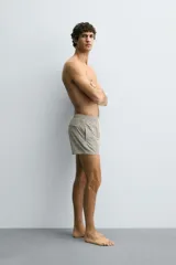 Bañador corto de hombre con cintura elástica ajustable mediante cordón blanco. Presenta un estampado geométrico repetitivo en tonos oscuros (negro/marrón oscuro) y blanco/beige. Incluye bolsillos laterales y un bolsillo trasero de vivo.