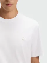 Remera de corte regular, color blanco, con cuello redondo y detalle de logo metálico circular en el pecho.