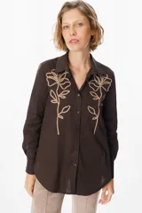 Camisa blanca de corte clásico con cuello camisero y cierre frontal de botones. Presenta un bordado lineal en color marrón oscuro en ambos lados del frente, representando motivos florales.