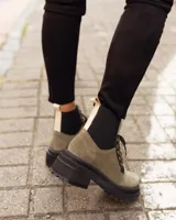 Borcego de gamuza color chocolate con paneles laterales elásticos negros y suela track dentada. Presenta sistema de cordones con ganchos metálicos y tirador en la parte trasera.