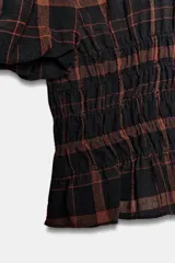 Camisa de cuello redondo y manga corta abullonada, con diseño de cuadros en tonos negro y marrón. Presenta un cuerpo entallado mediante frunces elásticos en la cintura y cierre posterior con abertura y botón.