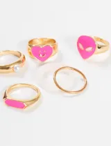 Set de anillos dorados con apliques de plástico rosa y blanco. Incluye un anillo con forma de corazón sonriente, un anillo con forma de alien, un anillo con aplique rectangular, un anillo con perla y dos anillos lisos.