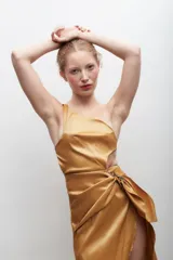 Vestido largo dorado de silueta al cuerpo, con escote asimétrico de un solo hombro, corte en la cintura con drapeado envolvente y lazo lateral, y falda con caída fluida y tajo frontal pronunciado.
