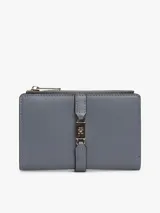 Billetera bifold Tommy Hilfiger de diseño compacto en color gris. Presenta un cierre superior con cremallera y un detalle frontal decorativo con broche metálico que incluye el logo de la marca. Cuenta con múltiples compartimentos internos para tarjetas y billetes.