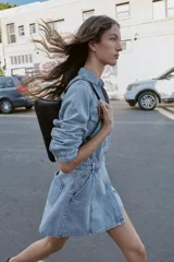 Vestido corto de denim azul claro, estilo camisa, con cuello solapa, escote pico, manga larga y cierre frontal de botones metálicos. Presenta bolsillos delanteros con tapa y bolsillos de plastrón en la espalda. La cintura está ajustada con elástico fruncido.