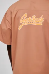 Sobrecamisa unisex color marrón claro, de manga corta y corte ancho. Presenta abertura frontal con botones tono sobre tono, dos bolsillos de parche en la parte inferior delantera y un bolsillo de ribete en el pecho. Logotipo de la marca estampado en contraste en el pecho y estampado grande en dos tonos en la espalda.