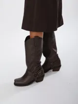 Bota estilo tejana de caña alta, confeccionada en cuero marrón con acabado texturizado. Presenta punta cuadrada, taco bajo y una correa con hebilla metálica ajustable y estribo desmontable en la zona del tobillo.