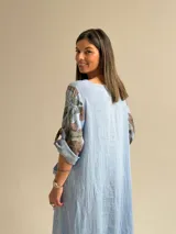 Vestido largo celeste con gran estampado floral marrón en el lateral derecho, de corte holgado, mangas 3/4 con trabilla y dobladillo irregular con flecos desflecados. Incluye un bolsillo aplicado en el frente.