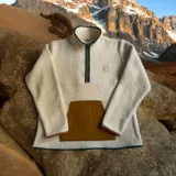 Buzo de polar color crema con cierre de medio largo con ribete verde, bolsillo marrón en el pecho y logo de la marca en el pecho.