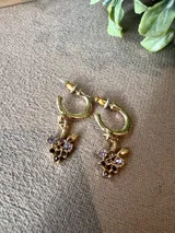 Aros tipo argolla pequeña en tono dorado con dije colgante en forma de racimo de uvas, decorado con pequeñas piedras brillantes en tonos violeta y morado.