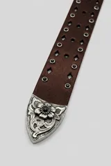 Cinto de cuero marrón con gran hebilla metálica plateada de estilo western, con grabados ornamentales, detalles de tachas y apliques circulares oscuros.