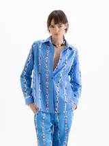 Camisa de calce holgado color azul, confeccionada en poplin satinado con estampado de motivos florales verticales. Presenta cuello clásico, mangas largas con puños abotonados y cierre frontal con botones.